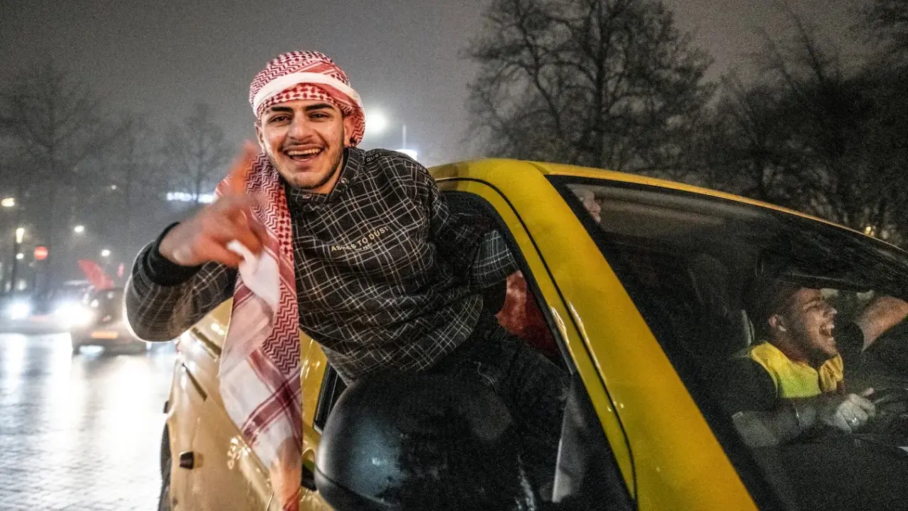 Jonge Marokkaanse Nederlanders vieren feest in een gele auto, met een van hen die uit het raam hangt en lachend zwaait.