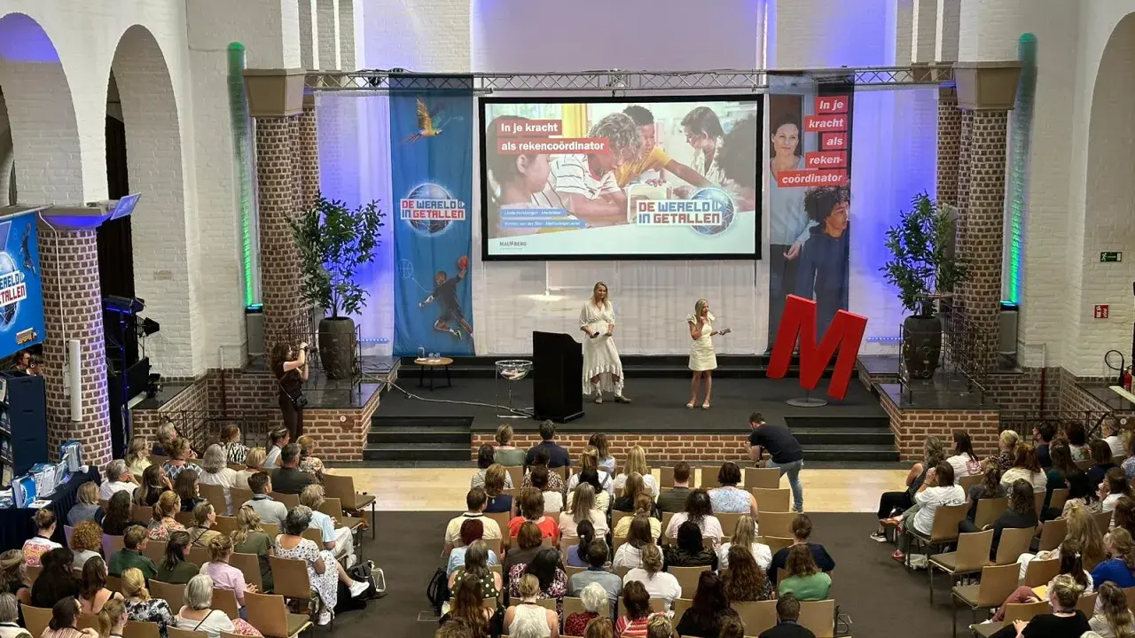 Publiek luistert naar presentatie over "de wereld in getallen". Twee sprekers op podium, met projectiescherm en grote rode M.