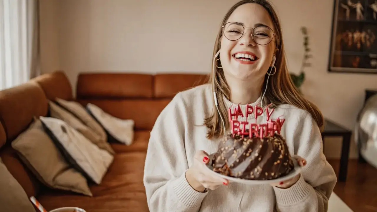 Vrolijke vrouw met bril en koptelefoon viert haar verjaardag met een chocoladetaart. Ze heeft de volwassen leeftijd bereikt en straalt van geluk.