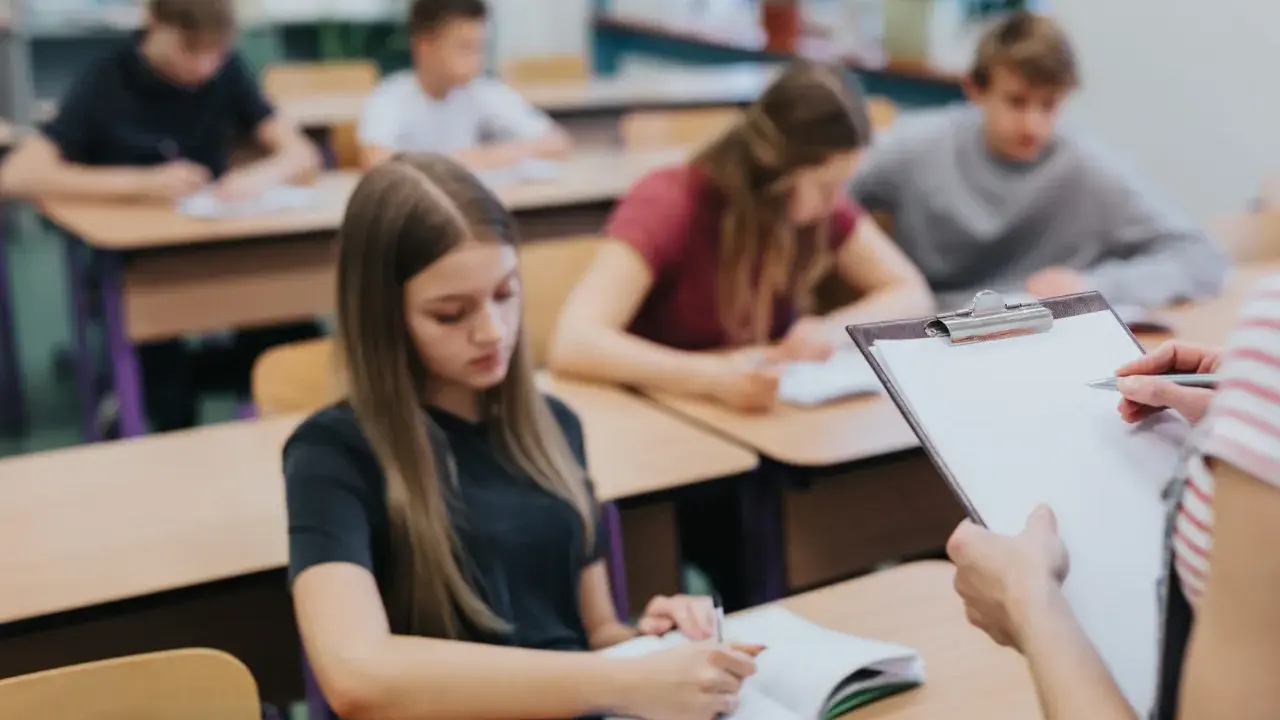 Leerlingen in een klaslokaal, sommigen schrijven in hun schrift. De leraar houdt een clipboard vast, misschien om te zien wat zijn leerlingen leren over wat zijn beta vakken.