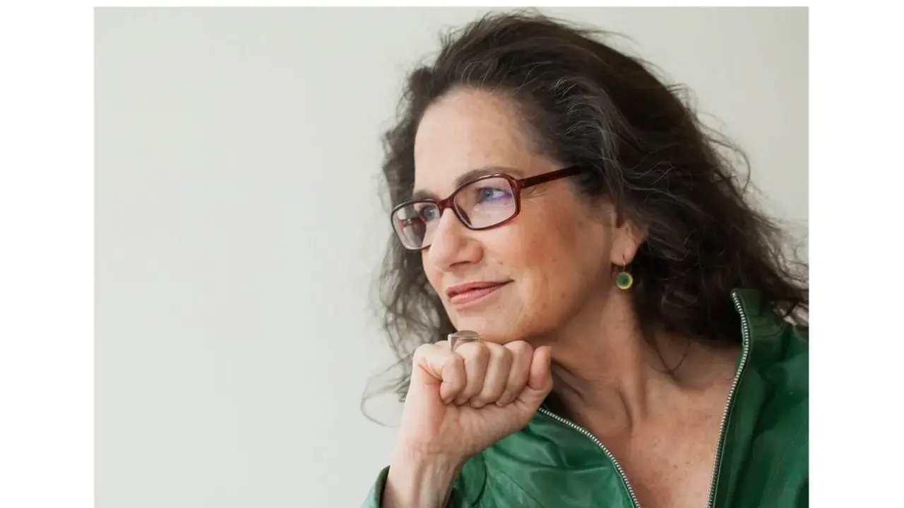 Susan Neiman, met een denkbeeldige blik, draagt een groene jas en een bril.