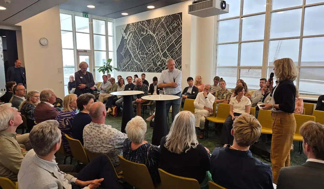 Mannen en vrouwen luisteren aandachtig naar een spreker, mogelijk over ruimtelijke ordening, in een zaal met een grote stadskaart aan de muur.