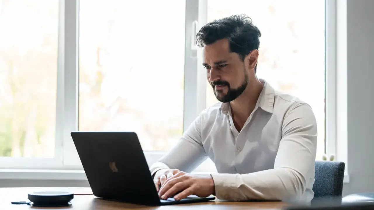 Man met baard, gekleed in een wit overhemd, geconcentreerd op zijn werk achter een laptop.