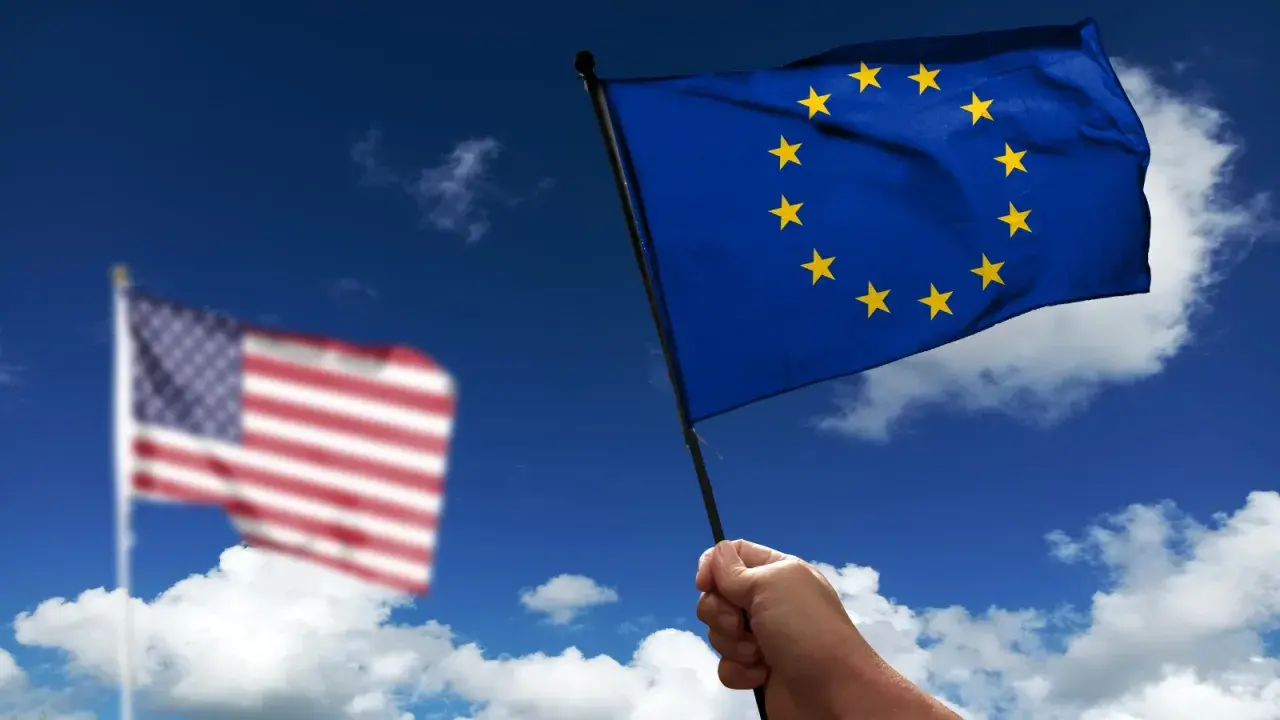 Hand holds EU flag, US flag blurred in background. Discussing what is soevereiniteit in a global context.