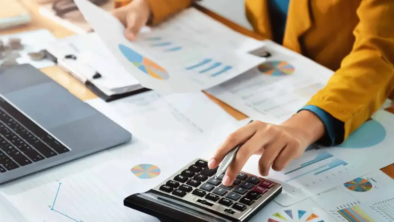 Persoon berekent cijfers op een calculator, omringd door grafieken en documenten. Dit kan te maken hebben met de commissie sociaal minimum.