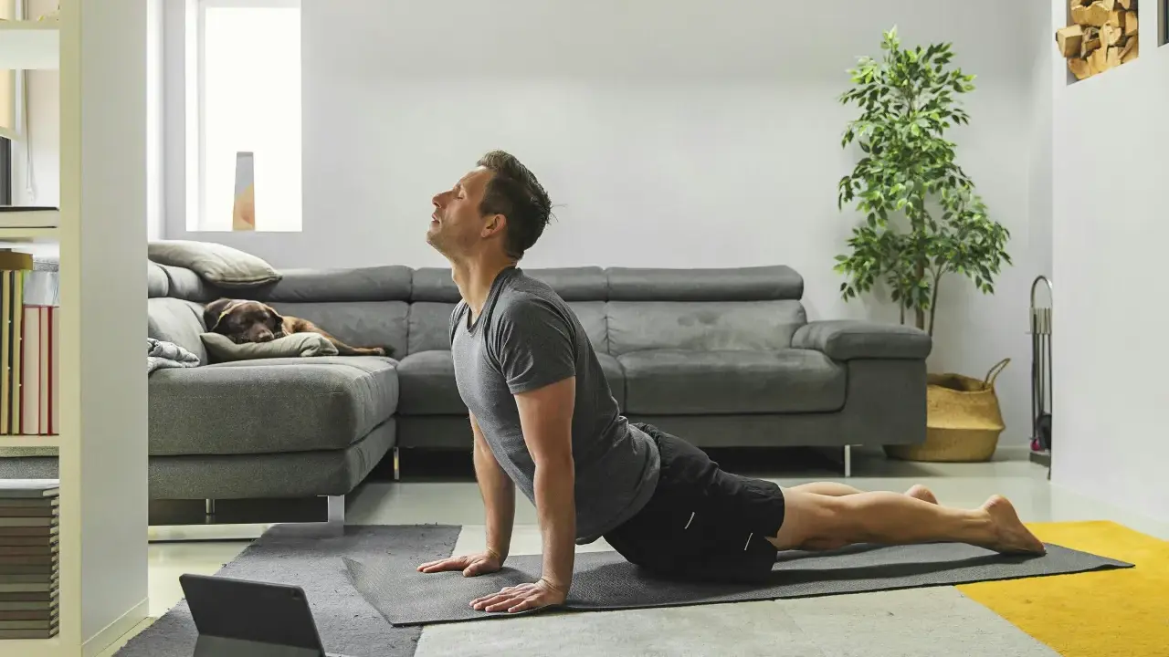Man in cobra pose op yogamat, neemt regie over zijn leven. Hond slaapt op bank.