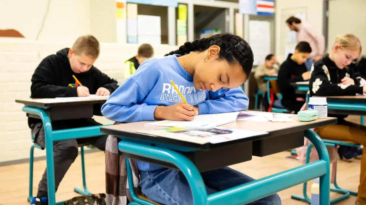 Kinderen maken hun werk, waarbij het leren zichtbaar wordt op papier. Een meisje met een blauwe trui schrijft geconcentreerd.