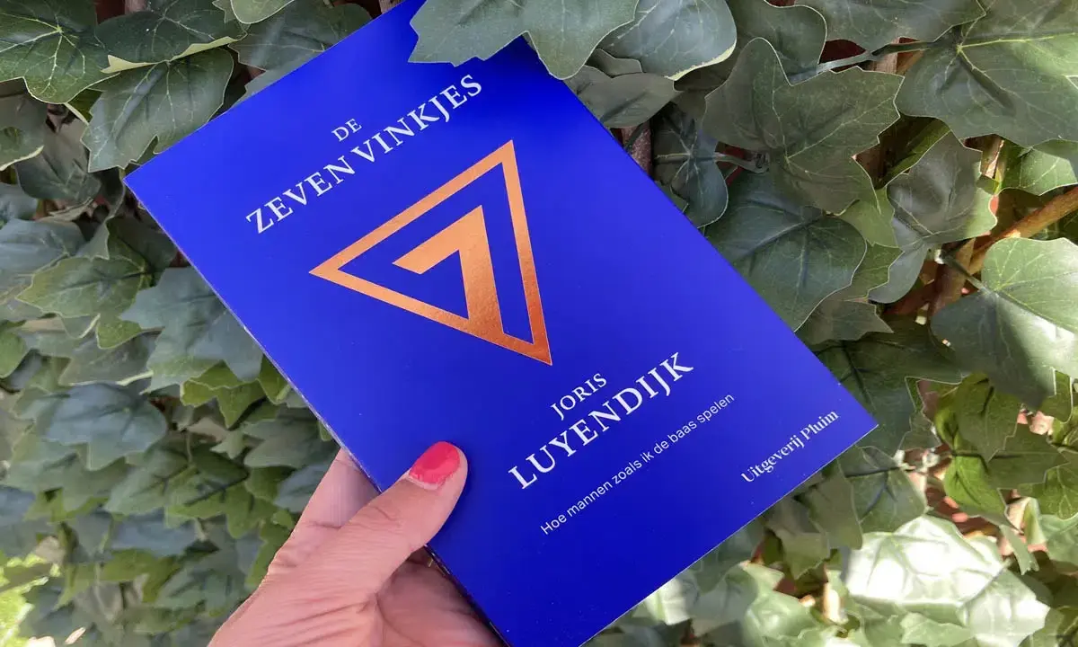Een hand houdt het boek "De Zeven Vinkjes" van Joris Luyendijk vast, omringd door groen blad.