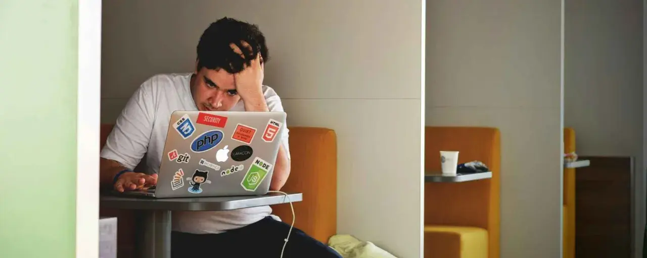 Een gefrustreerde jongere met zijn hand in zijn haar, starend naar zijn laptop vol met programmeerstickers. De druk van jongeren en schulden kan voelen als een eindeloze code.