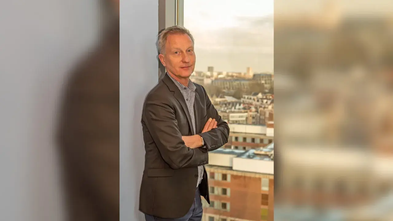 Jan Willem Duyvendak, een man met een grijze blazer en een geruit overhemd, staat met gekruiste armen voor een raam met uitzicht op een stad.