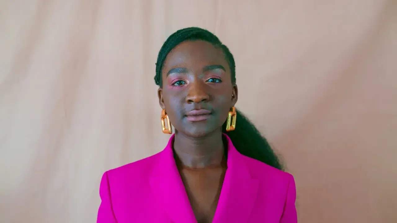 Een jonge vrouw van kleur met opvallende gouden oorbellen en een fuchsia blazer, kijkt zelfverzekerd de camera in.