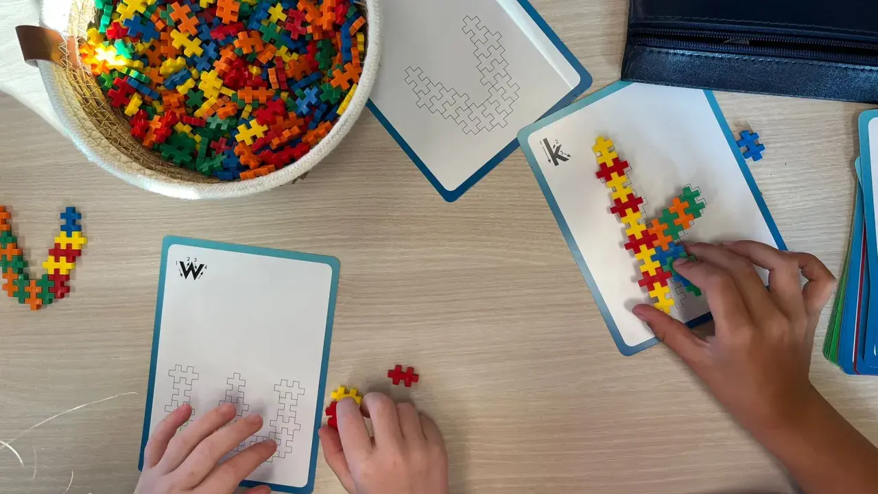 Kinderen bouwen met kleurrijke stukjes aan patronen op kaarten. Een mand vol bouwstenen ligt klaar voor meer taal spelletjes.