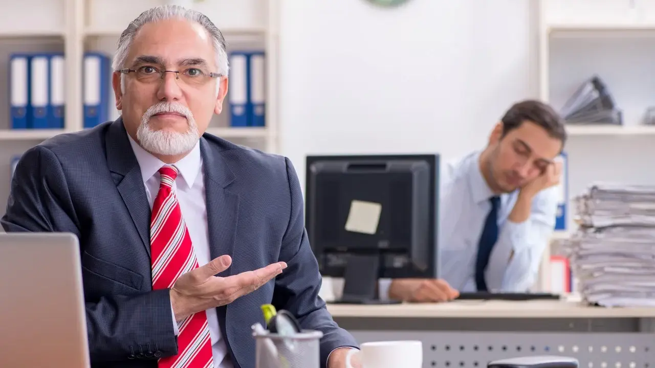 Een oudere man in pak legt iets uit, terwijl een jongere collega achter hem in slaap valt. Een beeld van volwassen worden en de uitdagingen van het werk.