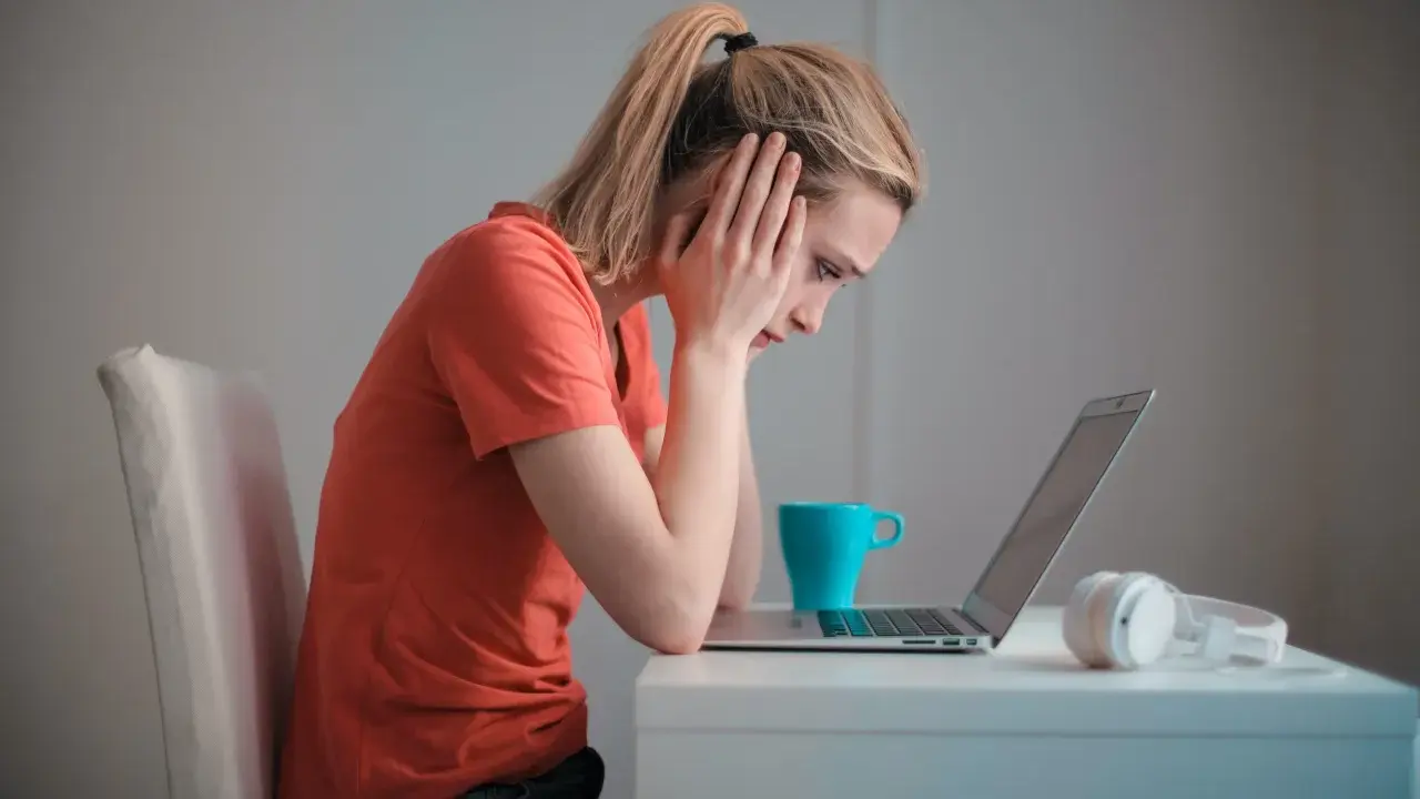 Een meisje met een korte spanningsboog staart naar haar laptop, handen aan haar hoofd. Een blauwe mok en koptelefoon staan naast haar.