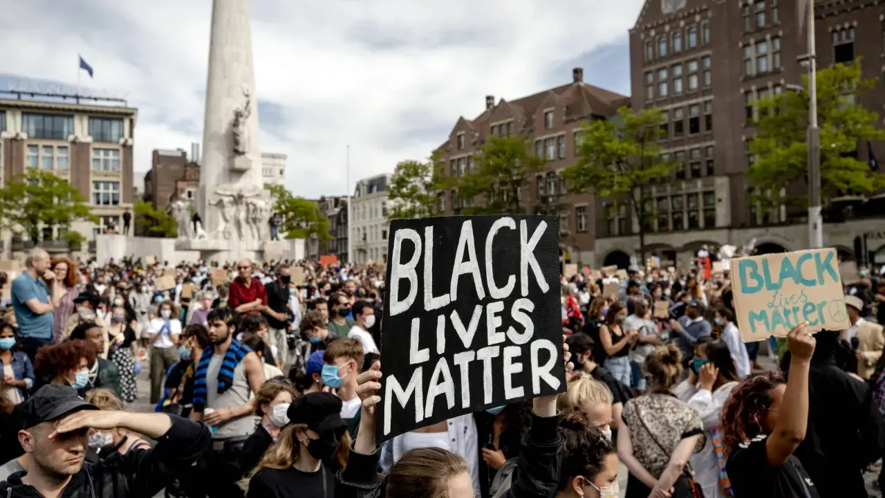 Mensen demonstreren op een plein met een monument, sommigen dragen borden met "Black Lives Matter".