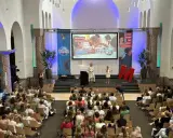 Publiek luistert naar presentatie over "de wereld in getallen". Twee sprekers op podium, met projectiescherm en grote rode M.
