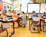 Een klas met kinderen die geconcentreerd aan hun tafels zitten. Een juf helpt een leerling, terwijl anderen werken op tablets of met potloden.