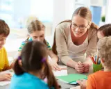 Docente begeleidt kinderen bij creatieve check-in werkvormen onderwijs. Kleurrijke potloden en notitieboekjes op tafel.