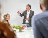 Een man spreekt enthousiast in een vergaderruimte, terwijl een vrouw aandachtig luistert. Ze vormen samen een sterk team.