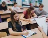 Leerlingen in een klaslokaal, sommigen schrijven in hun schrift. De leraar houdt een clipboard vast, misschien om te zien wat zijn leerlingen leren over wat zijn beta vakken.