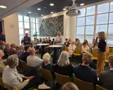 Mannen en vrouwen luisteren aandachtig naar een spreker, mogelijk over ruimtelijke ordening, in een zaal met een grote stadskaart aan de muur.