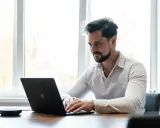 Man met baard, gekleed in een wit overhemd, geconcentreerd op zijn werk achter een laptop.