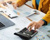 Persoon berekent cijfers op een calculator, omringd door grafieken en documenten. Dit kan te maken hebben met de commissie sociaal minimum.