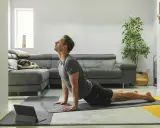 Man in cobra pose op yogamat, neemt regie over zijn leven. Hond slaapt op bank.