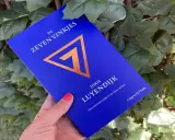 Een hand houdt het boek "De Zeven Vinkjes" van Joris Luyendijk vast, omringd door groen blad.