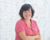 Een lachende vrouw met blauwe ogen, gekleed in een roze blouse, staat voor een witte bakstenen muur. Ze straalt de warmte uit van een zorgzame buurt.