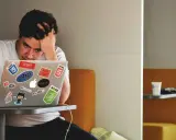 Een gefrustreerde jongere met zijn hand in zijn haar, starend naar zijn laptop vol met programmeerstickers. De druk van jongeren en schulden kan voelen als een eindeloze code.
