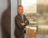 Jan Willem Duyvendak, een man met een grijze blazer en een geruit overhemd, staat met gekruiste armen voor een raam met uitzicht op een stad.