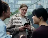 Drie vrouwen praten, één kijkt bezorgd naar haar telefoon met berichten als "Waar ben je?". Dit kan duiden op de angst die gepaard gaat met huiselijk geweld bij vrouwen.