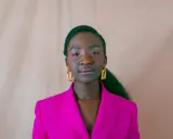 Een jonge vrouw van kleur met opvallende gouden oorbellen en een fuchsia blazer, kijkt zelfverzekerd de camera in.