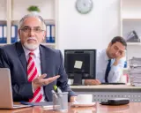 Een oudere man in pak legt iets uit, terwijl een jongere collega achter hem in slaap valt. Een beeld van volwassen worden en de uitdagingen van het werk.