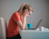 Een meisje met een korte spanningsboog staart naar haar laptop, handen aan haar hoofd. Een blauwe mok en koptelefoon staan naast haar.