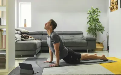 Man in cobra pose op yogamat, neemt regie over zijn leven. Hond slaapt op bank.