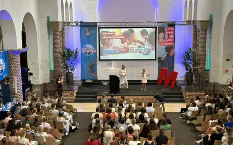 Publiek luistert naar presentatie over "de wereld in getallen". Twee sprekers op podium, met projectiescherm en grote rode M.