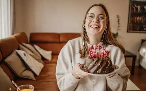 Vrolijke vrouw met bril en koptelefoon viert haar verjaardag met een chocoladetaart. Ze heeft de volwassen leeftijd bereikt en straalt van geluk.
