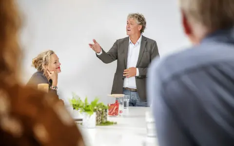 Een man spreekt enthousiast in een vergaderruimte, terwijl een vrouw aandachtig luistert. Ze vormen samen een sterk team.