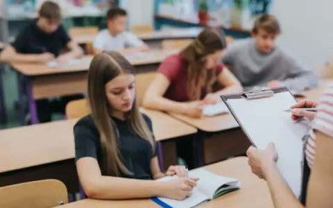 Leerlingen in een klaslokaal, sommigen schrijven in hun schrift. De leraar houdt een clipboard vast, misschien om te zien wat zijn leerlingen leren over wat zijn beta vakken.