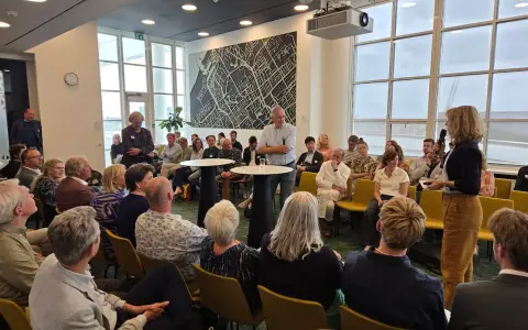 Mannen en vrouwen luisteren aandachtig naar een spreker, mogelijk over ruimtelijke ordening, in een zaal met een grote stadskaart aan de muur.