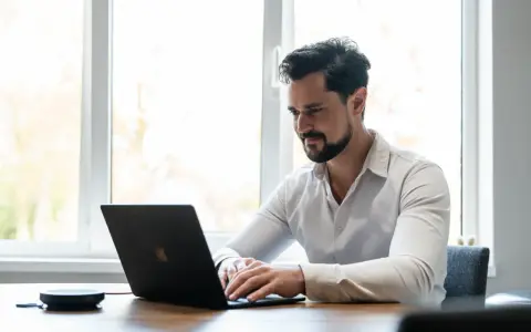 Man met baard, gekleed in een wit overhemd, geconcentreerd op zijn werk achter een laptop.