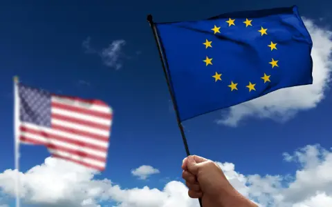 Hand holds EU flag, US flag blurred in background. Discussing what is soevereiniteit in a global context.