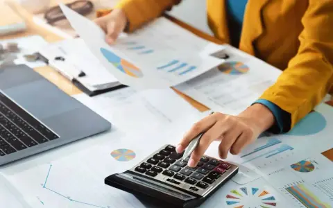 Persoon berekent cijfers op een calculator, omringd door grafieken en documenten. Dit kan te maken hebben met de commissie sociaal minimum.