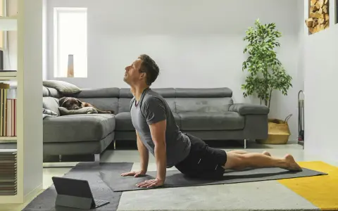Man in cobra pose op yogamat, neemt regie over zijn leven. Hond slaapt op bank.