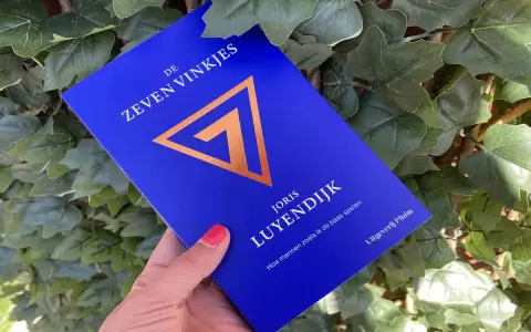 Een hand houdt het boek "De Zeven Vinkjes" van Joris Luyendijk vast, omringd door groen blad.