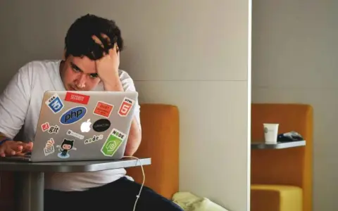 Een gefrustreerde jongere met zijn hand in zijn haar, starend naar zijn laptop vol met programmeerstickers. De druk van jongeren en schulden kan voelen als een eindeloze code.