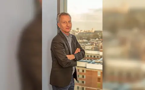 Jan Willem Duyvendak, een man met een grijze blazer en een geruit overhemd, staat met gekruiste armen voor een raam met uitzicht op een stad.