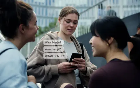 Drie vrouwen praten, één kijkt bezorgd naar haar telefoon met berichten als "Waar ben je?". Dit kan duiden op de angst die gepaard gaat met huiselijk geweld bij vrouwen.